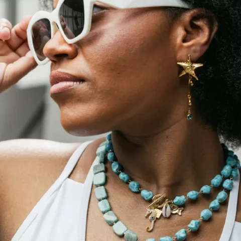 Μια χαρούμενη γυναίκα με σκούρα, σγουρά μαλλιά φοράει ένα λευκό αμάνικο τοπ και κομψά γυαλιά ηλίου με λευκό σκελετό. Χαμογελά πλατιά και κρατά το ένα χέρι ψηλά, ενισχύοντας την χαλαρή και ευτυχισμένη της διάθεση. Γύρω από το λαιμό της, φοράει ένα ζωντανό κολιέ με εναλλασσόμενες ογκώδεις χάντρες τυρκουάζ και αμαζονίτη, που διαθέτει ένα μικρό χρυσό κέλυφος στο κέντρο. Φοράει επίσης ένα χρυσό σκουλαρίκι σε σχήμα αστερία, προσθέτοντας μια θαλασσινή έμπνευση στην εμφάνισή της. Το φωτεινό φόντο τονίζει την χαρούμενη έκφρασή της και τα ζωηρά χρώματα των αξεσουάρ της.