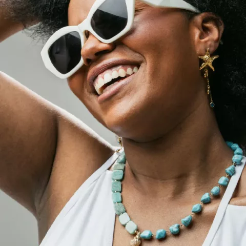Μια χαρούμενη γυναίκα με σκούρα, σγουρά μαλλιά φοράει ένα λευκό αμάνικο τοπ και κομψά γυαλιά ηλίου με λευκό σκελετό. Χαμογελά πλατιά και κρατά το ένα χέρι ψηλά, ενισχύοντας την χαλαρή και ευτυχισμένη της διάθεση. Γύρω από το λαιμό της, φοράει ένα ζωντανό κολιέ με εναλλασσόμενες ογκώδεις χάντρες τυρκουάζ και αμαζονίτη, που διαθέτει ένα μικρό χρυσό κέλυφος στο κέντρο. Φοράει επίσης ένα χρυσό σκουλαρίκι σε σχήμα αστερία, προσθέτοντας μια θαλασσινή έμπνευση στην εμφάνισή της. Το φωτεινό φόντο τονίζει την χαρούμενη έκφρασή της και τα ζωηρά χρώματα των αξεσουάρ της.
