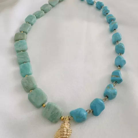 Ένα κομψό κολιέ διαθέτει εναλλασσόμενες ογκώδεις χάντρες τυρκουάζ και αμαζονίτη, συνδεδεμένες μεταξύ τους με μικρούς χρυσούς συνδέσμους. Το κεντρικό στοιχείο είναι ένα κοχύλι, είναι ένα περίτεχνο διακοσμημένο χρυσό κέλυφος που κρέμεται από το κέντρο του κολιέ. Το κομμάτι εκτίθεται σε ένα απαλό λευκό ύφασμα, αναδεικνύοντας τον σχεδιασμό με έμπνευση από την ακτή και τις ζωντανές γαλαζοπράσινες αποχρώσεις του.